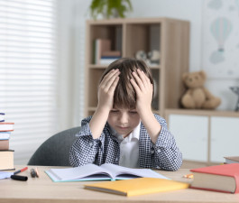 Pamatskolas skolēns saķēris galvu sēž pie darba burtnīcas, uz galda ir grāmatas 